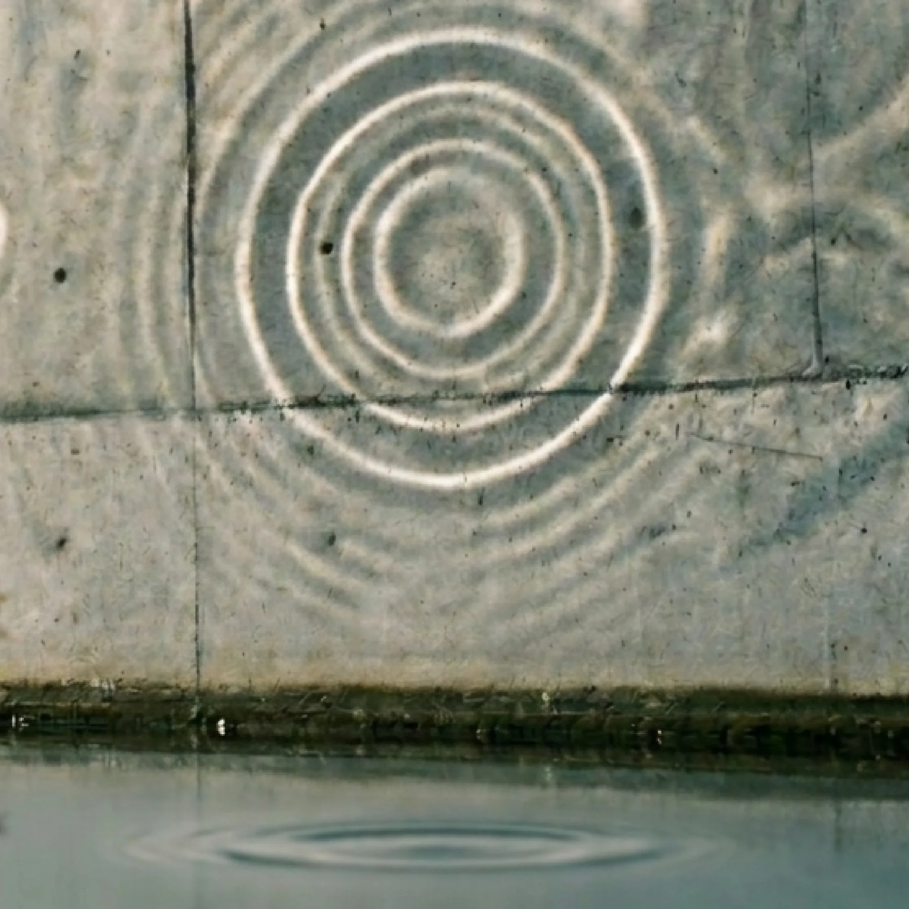 Rings of reflected light from rippling water thrown across a board-formed concrete wall, with the water's edge and a faint ripple below.