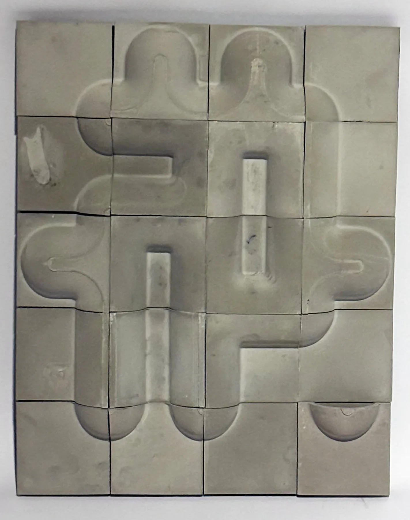 Cast concrete test panel of twenty square tiles mounted on a white wall, with rounded relief channels running continuously across the joints.