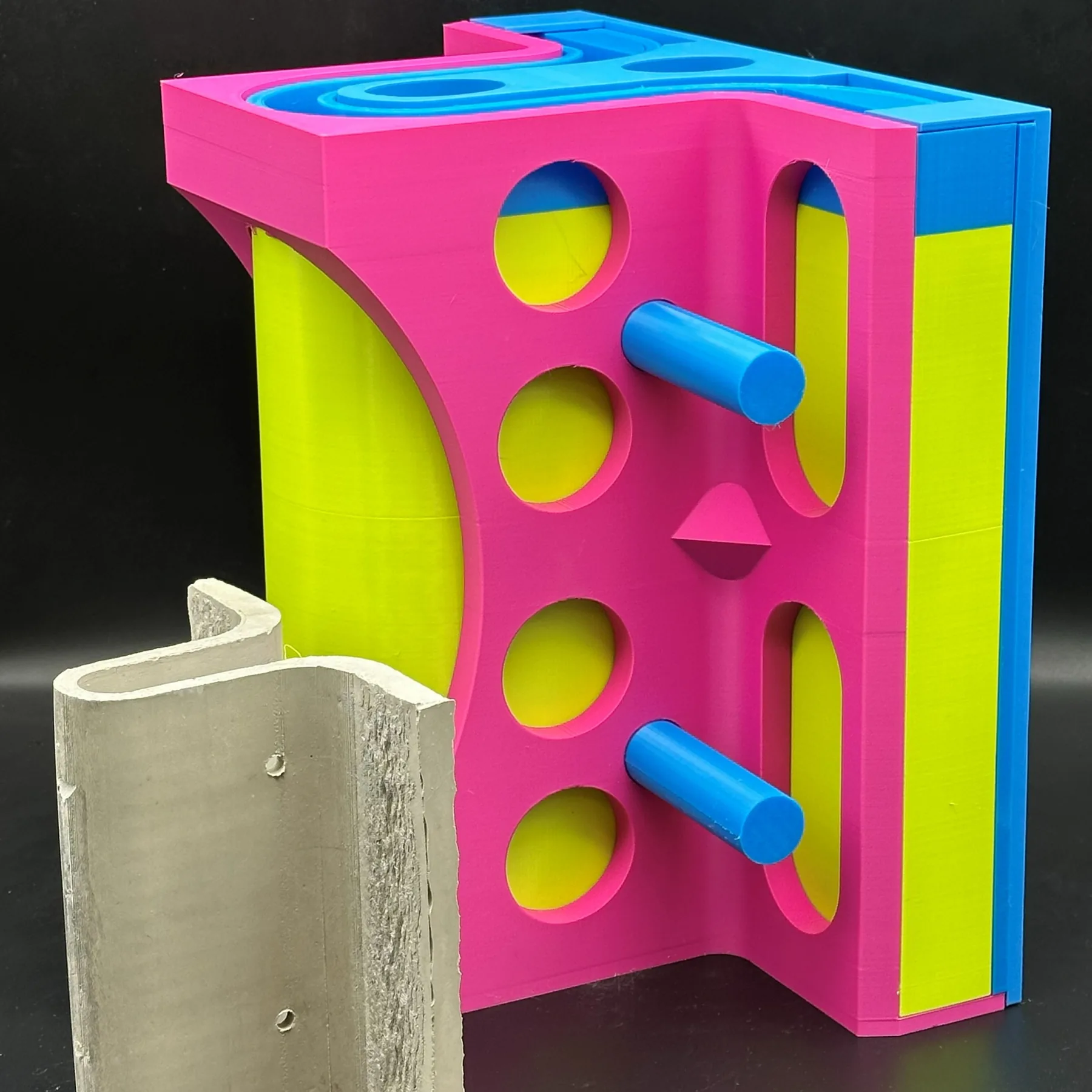 Assembled magenta, blue and lime-green 3D-printed mold with round and slotted windows, a demolded grey concrete pipe casting with pin holes standing in front.